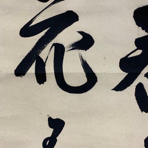 May include: Close-up of a section of a traditional Asian calligraphy artwork. Bold, black ink brushstrokes form stylized characters on a cream-colored paper background. The strokes vary in thickness, creating a dynamic visual texture. The artwork is likely part of a larger piece.