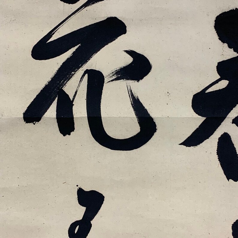 May include: Close-up of a section of a traditional Asian calligraphy artwork. Bold, black ink brushstrokes form stylized characters on a cream-colored paper background. The strokes vary in thickness, creating a dynamic visual texture. The artwork is likely part of a larger piece.