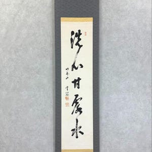 以下が含まれることがあります： クリーム色の背景に黒インクで書かれた書道が施された、縦長の日本の掛け軸。 軸は、テクスチャパターンを持つ濃い灰色の生地で縁取られています。 テキストは縦書きで、赤い印鑑が含まれています。