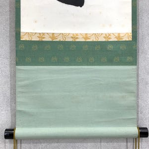 Puede incluir: Un rollo de pintura japonesa tradicional con un dise&ntilde;o de tinta negra sobre papel blanco. El rollo presenta un borde verde y dorado estampado y un fondo verde claro. El rollo est&aacute; enrollado con clavijas de madera negra y cordones dorados.