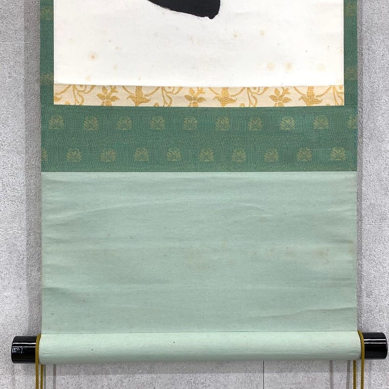 Puede incluir: Un rollo de pintura japonesa tradicional con un dise&ntilde;o de tinta negra sobre papel blanco. El rollo presenta un borde verde y dorado estampado y un fondo verde claro. El rollo est&aacute; enrollado con clavijas de madera negra y cordones dorados.