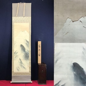May include: A long, vertical Japanese scroll painting of a mountain landscape in grey and white tones. The artwork is mounted on a cream background with a grey roller at the base. A wooden box with Japanese text is positioned beside the scroll.