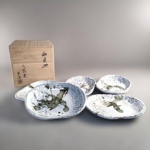 May include: A set of six white ceramic dishes with a textured surface and dark grey abstract designs. A wooden box with Japanese characters is in the background. The dishes are oval and round.