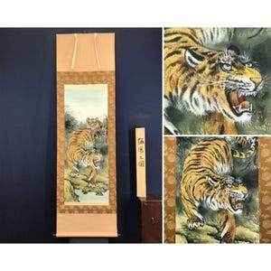 May include: A vertical hanging scroll featuring a painting of two tigers in a natural landscape. The artwork displays a large tiger with an open mouth and a smaller tiger behind it. The scroll has a tan border with a floral pattern and text.