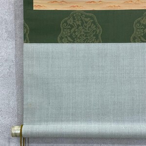 May include: A traditional Japanese scroll painting with a light blue fabric and a green patterned section. The top section features a beige border with a landscape design. The scroll is rolled up at the bottom, revealing a wooden dowel.