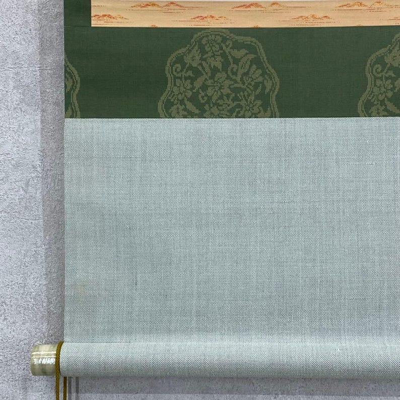 May include: A traditional Japanese scroll painting with a light blue fabric and a green patterned section. The top section features a beige border with a landscape design. The scroll is rolled up at the bottom, revealing a wooden dowel.