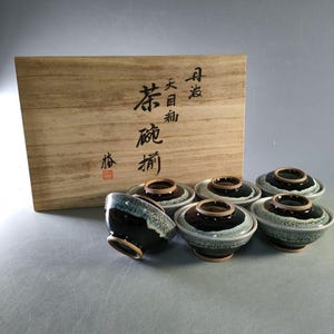 May include: A set of six ceramic tea bowls with lids, featuring a dark blue and white glaze. The bowls are arranged in front of a wooden box with Japanese calligraphy.