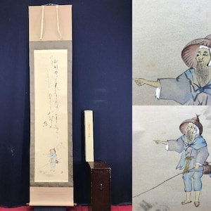 Op de afbeelding: Een antiek Japans rolschilderij met kalligrafie en een kleine illustratie van een vissersfiguur. De rol is crèmekleurig met een beige boven- en onderkant. Een kleine houten kist staat naast de rol.