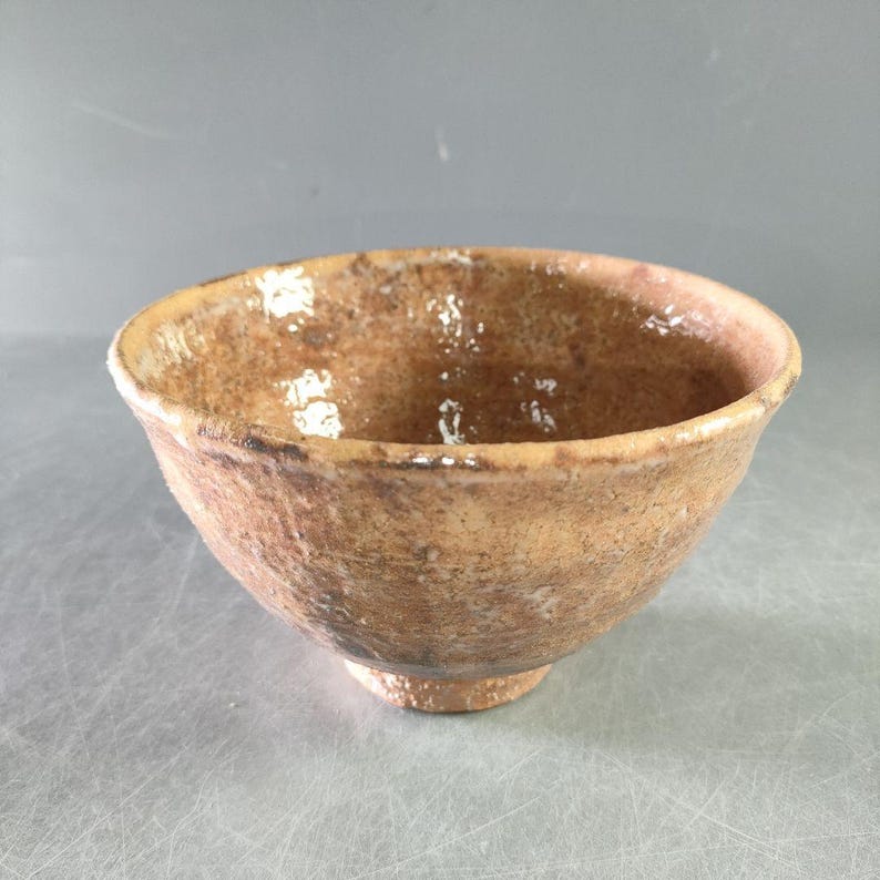 May include: A handmade, rustic ceramic bowl with a textured, earthy brown exterior. The bowl has a wide, open mouth and a slightly raised base. The interior has a glossy finish, and the bowl appears to be hand-thrown.