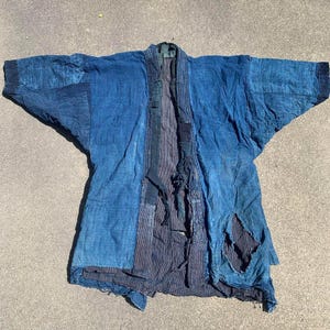 May include: A vintage indigo-dyed Japanese kimono jacket, with a patchwork design in various shades of blue. The jacket has wide sleeves and shows signs of wear, indicating its age. The garment is laid flat on a neutral surface.