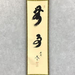 May include: A vertical scroll featuring Japanese calligraphy in black ink on a cream-colored background. The calligraphy includes two large characters and smaller text with red stamps. The scroll is framed with a gold border.