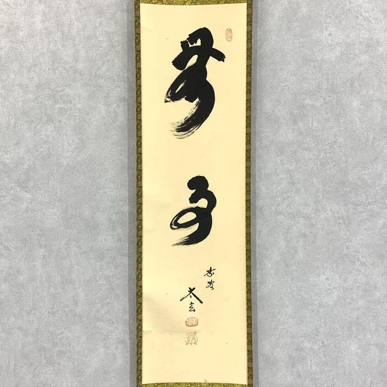 May include: A vertical scroll featuring Japanese calligraphy in black ink on a cream-colored background. The calligraphy includes two large characters and smaller text with red stamps. The scroll is framed with a gold border.