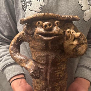 May include: A handcrafted, brown ceramic sculpture of a whimsical creature with a smiling face, arms, and a hat. The textured surface and earthy tones give it a rustic, artistic feel. The sculpture is held in front of a gray sweatshirt.