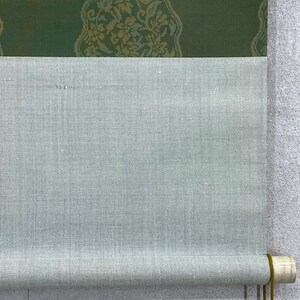 May include: A rolled-up scroll featuring a light green fabric with a textured surface. The top portion has a dark green panel with a gold floral pattern. The scroll is held together with a wooden dowel and a gold cord.