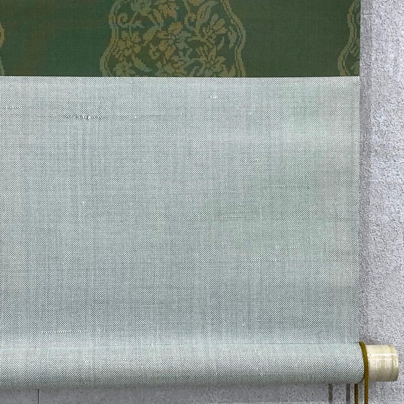May include: A rolled-up scroll featuring a light green fabric with a textured surface. The top portion has a dark green panel with a gold floral pattern. The scroll is held together with a wooden dowel and a gold cord.