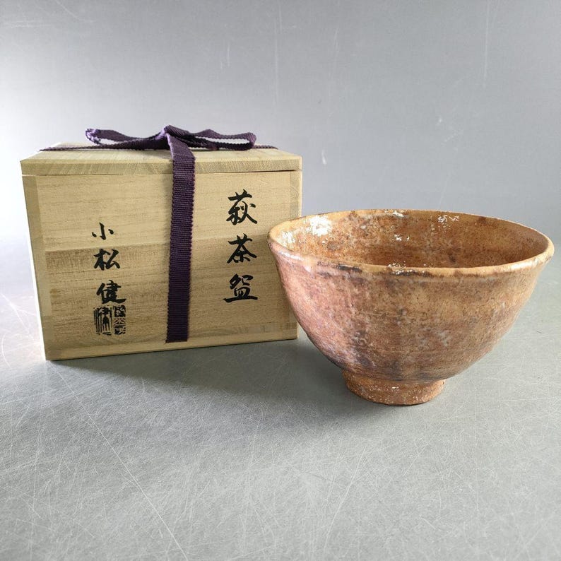 May include: A rustic, handcrafted tea bowl with a textured, earthy brown glaze. The bowl sits beside a wooden box tied with a purple ribbon. The box has Japanese characters and a stamp.