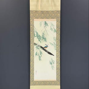 May include: A vertical hanging scroll featuring a painting of a small blue and orange kingfisher bird perched on a dark tree branch. Green willow branches hang down from the top. The painting is framed by patterned fabric borders in cream and grey.