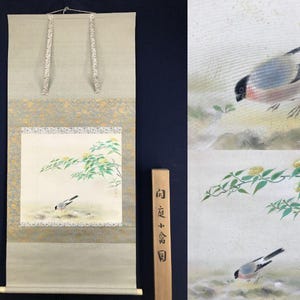 Könnte beinhalten: Eine traditionelle japanische Hängerolle mit einer Malerei eines Vogels in der Nähe eines blühenden Zweigs. Die Rolle hat einen hellen Hintergrund mit goldenen Akzenten und eine hölzerne Aufbewahrungsbox. Das Kunstwerk ist im traditionellen japanischen Stil.
