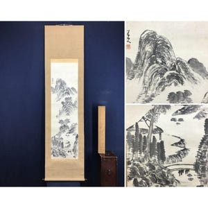 May include: An Asian-style ink painting on a scroll, depicting a mountainous landscape with trees and a small structure. The artwork is primarily in shades of black and white, with a neutral-toned scroll backing. A wooden box and a wooden stick are next to the scroll.