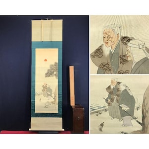 Op de afbeelding: Een antiek Japans rolschilderij dat een serene winterlandschap met figuren uitbeeldt. Het kunstwerk toont een felrode zon, een boom en figuren in traditionele kledij. De rol is omlijst met blauwe en groene stof en bevat een houten opbergdoos.