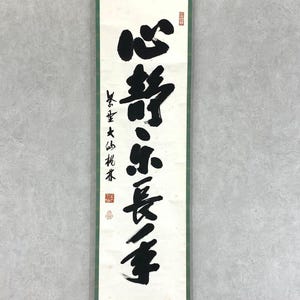 Puede incluir: Un pergamino vertical con caligraf&iacute;a negra sobre fondo blanco, enmarcado por un borde verde. La caligraf&iacute;a presenta trazos grandes y audaces de caracteres chinos. Sellos rojos son visibles en la parte superior e inferior, agregando un toque tradicional.