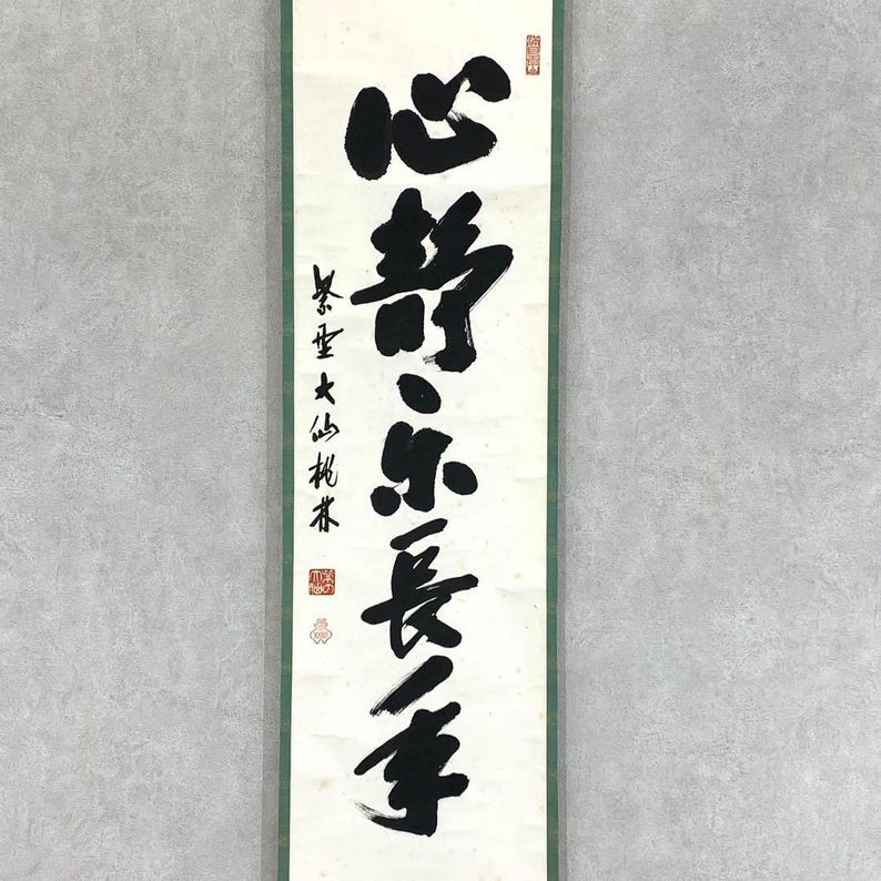 Puede incluir: Un pergamino vertical con caligraf&iacute;a negra sobre fondo blanco, enmarcado por un borde verde. La caligraf&iacute;a presenta trazos grandes y audaces de caracteres chinos. Sellos rojos son visibles en la parte superior e inferior, agregando un toque tradicional.