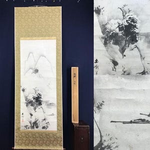Könnte beinhalten: Traditionelle japanische Rollbildmalerei mit einer monochromen Landschaft mit Bergen, Bäumen und einem Fluss. Das Kunstwerk ist von einem goldfarbenen, gemusterten Rand umgeben und enthält eine Holzscrollleiste. Die Malerei wird vor einem dunkelblauen Hintergrund präsentiert.