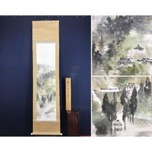 Op de afbeelding: Een traditioneel Japans rolschilderij dat een landschap met bergen, bomen en gebouwen afbeeldt. Het kunstwerk is voornamelijk in tinten grijs, groen en bruin, met een beige rand. Een houten kist en een houten stok met Japanse tekst staan naast de rol.