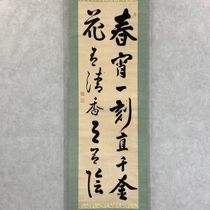 May include: A vertical scroll with black calligraphy on a cream-colored background. The text is in traditional Chinese characters. The scroll is framed by a green border and a gold decorative trim at the bottom. The calligraphy is the primary focus.