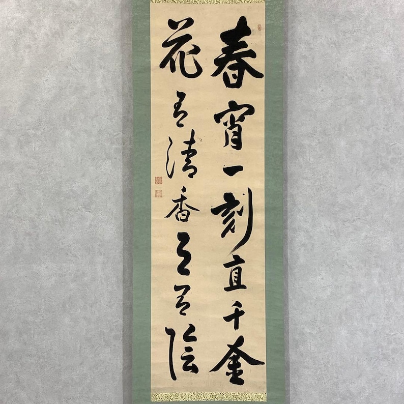 May include: A vertical scroll with black calligraphy on a cream-colored background. The text is in traditional Chinese characters. The scroll is framed by a green border and a gold decorative trim at the bottom. The calligraphy is the primary focus.