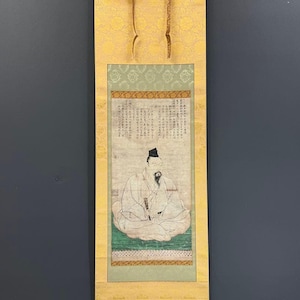 Op de afbeelding: Een verticale hangende rol met een centraal schilderij van een zittende figuur in traditionele kleding met een zwarte muts en baard. Het kunstwerk bevat kolommen met Japanse kalligrafie boven de figuur, omlijst door lichtgroene en gouden stoffen randen.