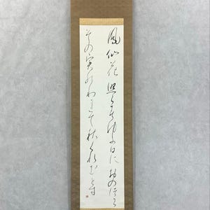 May include: A vertical Japanese scroll featuring black calligraphy on a white background. The scroll is framed by a brown patterned border and has a wooden rod at the bottom. The text is in Japanese.