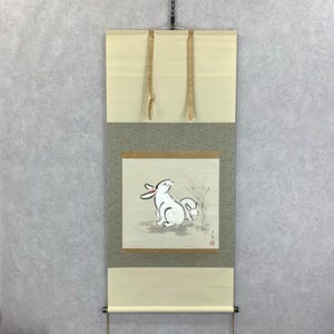May include: Japanese wall scroll with a watercolour painting of two white rabbits. The artwork is set against a neutral background with a cream top and bottom, and a grey-green middle section. The scroll is suspended by a black cord.