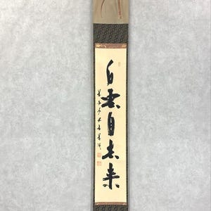May include: A vertical Japanese scroll with calligraphy in black ink on a cream-colored background. The scroll has a tan top and bottom with decorative patterned fabric and a metal bar at the bottom.