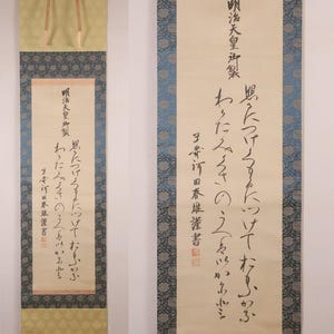 May include: Two Japanese calligraphy scrolls. The scrolls display black ink characters on a cream background, framed by patterned borders in blue and gold. Each scroll has a gold ribbon at the top for hanging. The text is in Japanese.