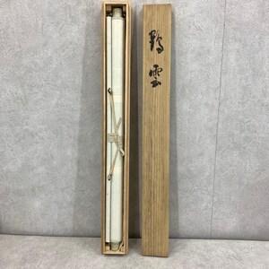 May include: A rolled scroll, secured with a beige cord, is housed in a rectangular wooden box. The box is open, revealing the scroll's off-white surface. A separate wooden lid with black Japanese characters is placed next to the box.