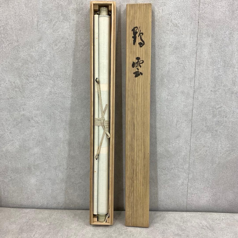 May include: A rolled scroll, secured with a beige cord, is housed in a rectangular wooden box. The box is open, revealing the scroll's off-white surface. A separate wooden lid with black Japanese characters is placed next to the box.