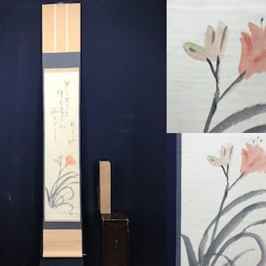 May include: A long, vertical scroll painting featuring a floral design with pink and white flowers and green leaves. The artwork includes Japanese calligraphy. The scroll is framed with blue and beige borders and is displayed against a dark blue background.