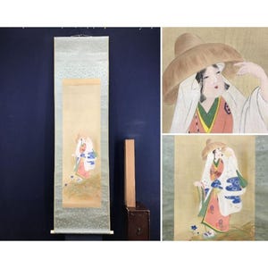 Op de afbeelding: Een Japanse hangrol met een schildering van een figuur met een breedgerande strooien hoed, een roze gewaad met groene rand en een wit bovenkleed met blauwe bloemmotieven. De figuur staat op een grasveldje met kleine blauwe bloemen.
