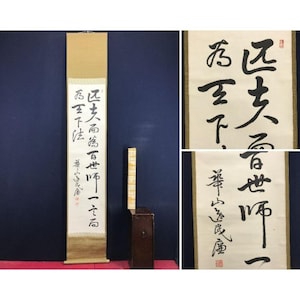 Op de afbeelding: Een traditionele Japanse rol met zwarte kalligrafie op een crèmekleurige achtergrond. De rol, met tekst en een rood zegel, wordt verticaal gepresenteerd. Een houten kist staat naast de rol.