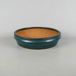 May include: A small, round, ceramic bowl with a teal exterior and a brown interior. The bowl has a slightly raised rim and a smooth, glossy finish. The bowl is suitable for serving small portions of food or as a decorative item.