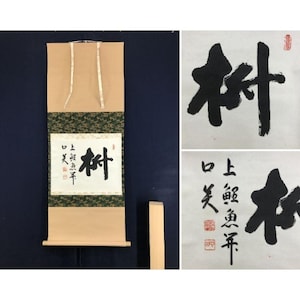 May include: Japanese calligraphy scroll featuring the character for "tree" in bold black ink. The scroll has a tan backing with a green and gold patterned border. Additional calligraphy examples are shown on white paper.