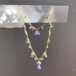 May include: A delicate gold chain necklace with multiple layers of green and purple flower charms. The necklace features small, clear beads and a dangling purple flower charm.
