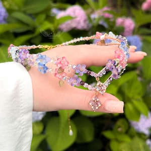 May include: A multi-strand choker necklace featuring delicate pink, blue, and purple glass flowers interspersed with small clear beads and gold-toned metal leaves. A central diamond-shaped pendant with pink crystals hangs from the floral arrangement.