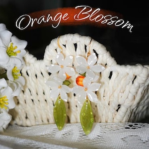 May include: White flower earrings with gold-colored hooks and green leaf-shaped dangles. The earrings are displayed in a woven basket. The image includes the text "Template for Shrink Plastic Creations Cutting Machine & Hand-Cut" and "Orange Blossom".