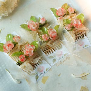 May include: Several hair combs adorned with delicate, pink and white rose-like flowers and green leaves. Each comb is embellished with a sheer, cream-colored ribbon and a small card with the text "lavila jewelry".