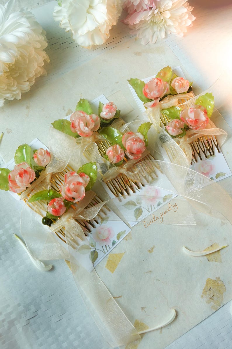 May include: Several hair combs adorned with delicate, pink and white rose-like flowers and green leaves. Each comb is embellished with a sheer, cream-colored ribbon and a small card with the text "lavila jewelry".