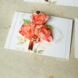 May include: A floral hair clip featuring three coral-colored resin roses with green leaves. The clip has a gold-colored rectangular base with a bee charm. The hair clip is displayed on a white card with the text "Hair Clip" and "lavila jewelry".