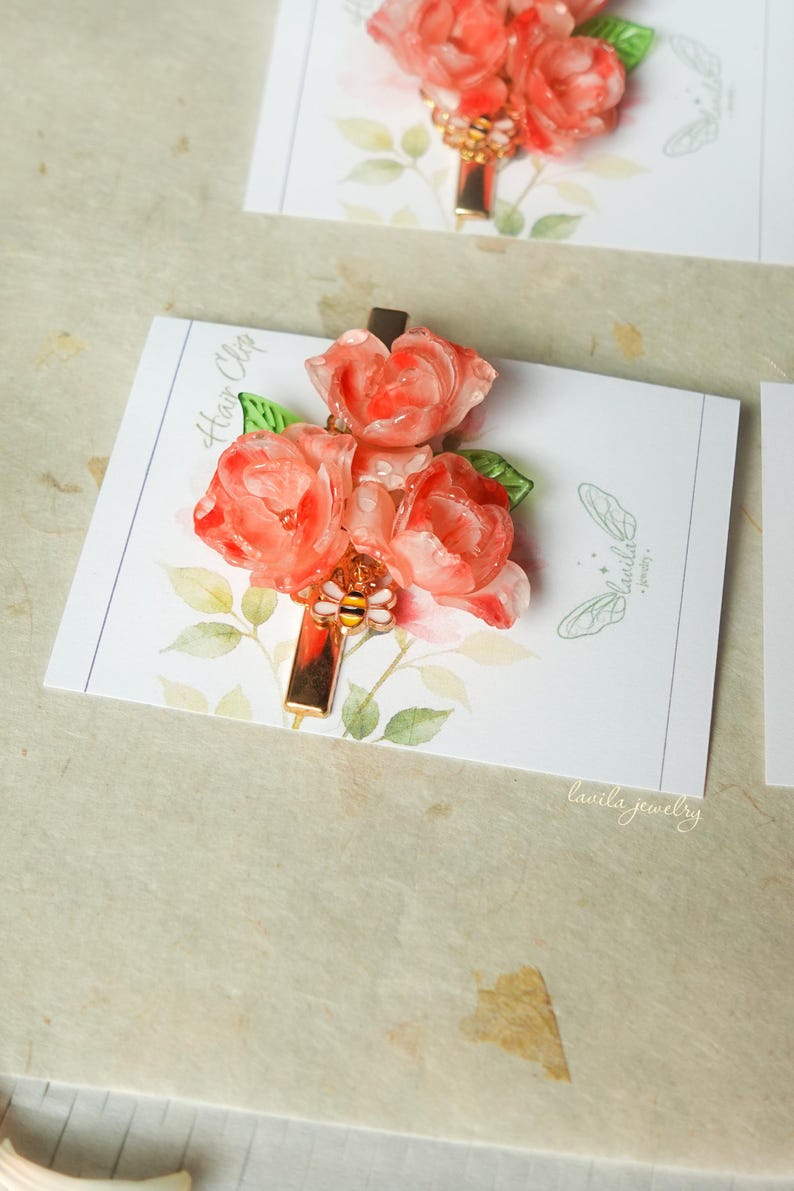 May include: A floral hair clip featuring three coral-colored resin roses with green leaves. The clip has a gold-colored rectangular base with a bee charm. The hair clip is displayed on a white card with the text "Hair Clip" and "lavila jewelry".