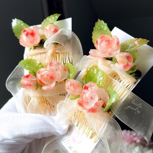 May include: Two floral hair combs with pink rose accents and green leaves. The combs have gold-colored teeth and are adorned with sheer ribbons. The text "lavila jewelry" is visible on one of the ribbons.
